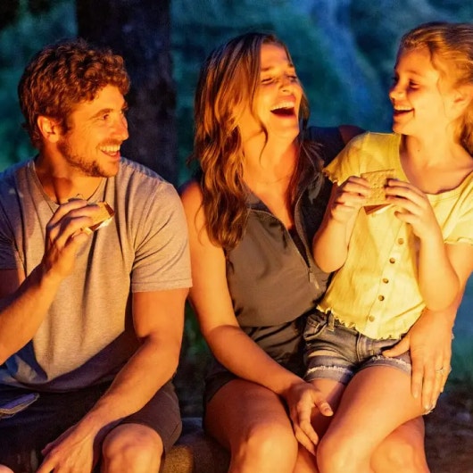 family sitting around fire and smiling while eating smores